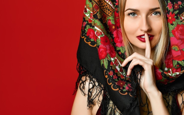 Shot of beautiful female with long blonde hair wears kokoshnik cap and patterned shawl shows hush sign