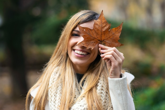 Shot of beautiful smiling woman having fun with leave while covering his eye with it looking at the camera in the park in autumn.