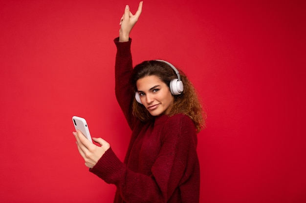 Shot of beautiful young brunette curly woman wearing dark red sweater isolated over red background