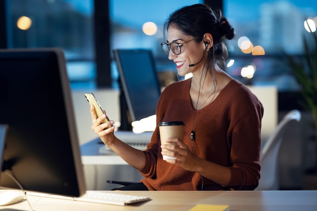 Shot of beautiful young business woman having videocall with mobile phone while working with computer sitting in the office.