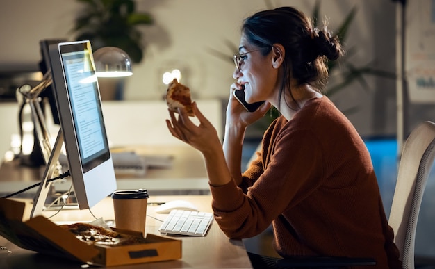 Shot of beautiful young business woman working with computer while eating pizza sitting in the office.