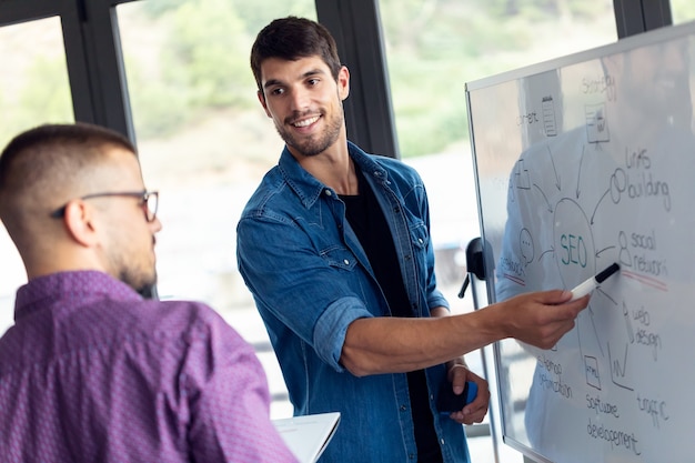 Shot of business young man pointing on whiteboard while discussing with his colleague of they new project in the modern startup office.