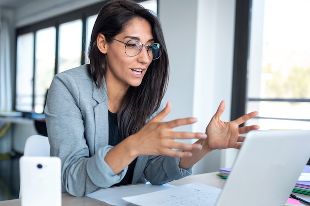 Shot of confident business woman looking and speaking through the webcam while making a video conference with laptop from the office.