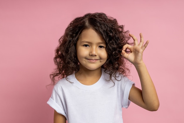 Shot of confident pretty little caucasian girl with dark curly hair, in white t shirt, raising hand and showing ok sign, expressing approval. Gesture, body language.