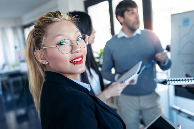 Shot of elegant young businesswoman looking at camera while her colleagues are explaining to the project in coworking place.