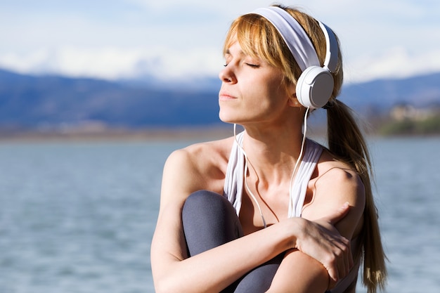 Shot of fit and sporty young woman relaxing and listening to music after work out next to the lake.