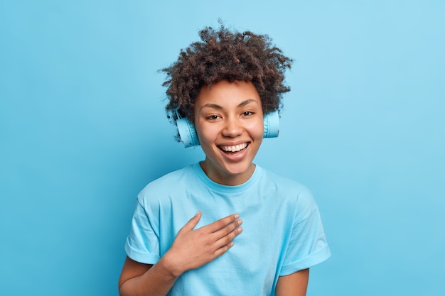  shot of glad Afro American girl smiles positively dressed in casual t shirt listes music via headphones being amused poses against blue wall. Monochrome shot. Positive emotions.