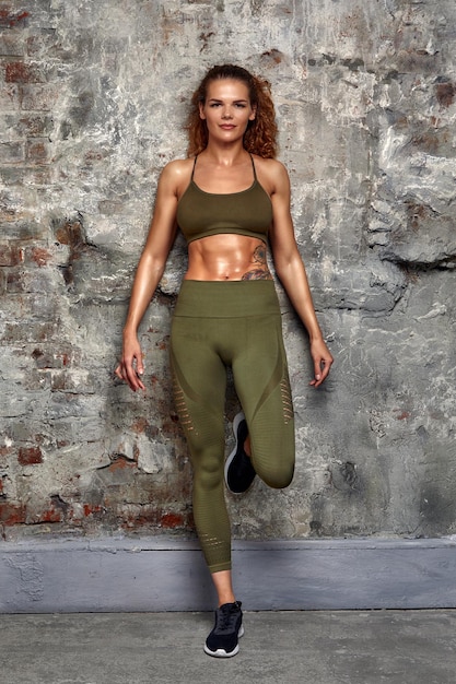 Shot of good looking sporty slim woman dressed in sportsclothes with muscular abs has active lifestyle poses against grey concrete background Lifestyle concept