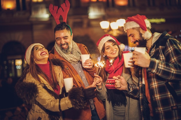 Shot of a group of cheerful young friends having fun with sparklers  and enjoying hot drink on the Christmas market at an evening party.