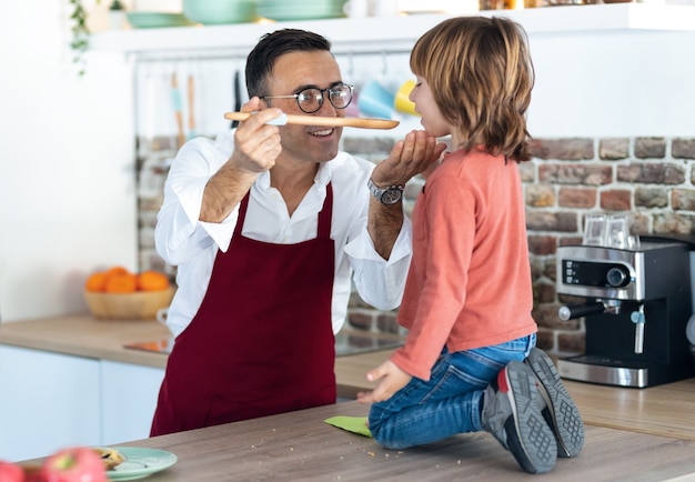 Shot of handsome father cooking and letting his son taste the food in the kitchen at home.