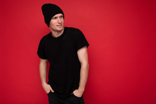 Shot of handsome positive young male person standing isolated over red background wall wearing black t-shirt for mockup and black hat and looking to the side