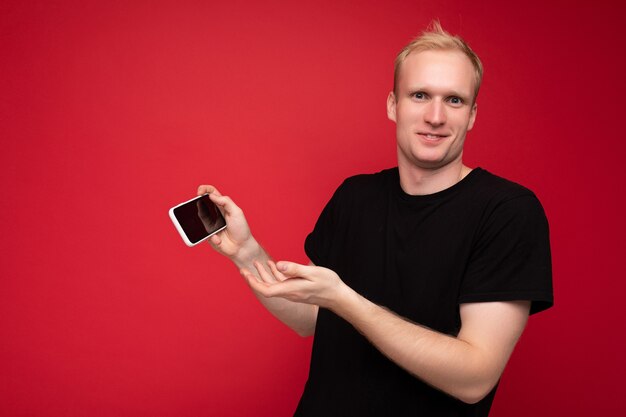 Shot of handsome smiling blonde young male person wearing black t-shirt standing isolated on red background with copy space holding smartphone showing phone in hand with empty display for mockup point