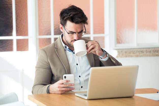 Shot of handsome young business man using his smartphone while drinking coffee in front of laptop in the office.