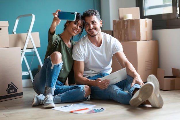 Shot of lovely couple taking a picture of themselves while looking ideas to decorate the new apartment in digital tablet sitting on the floor.