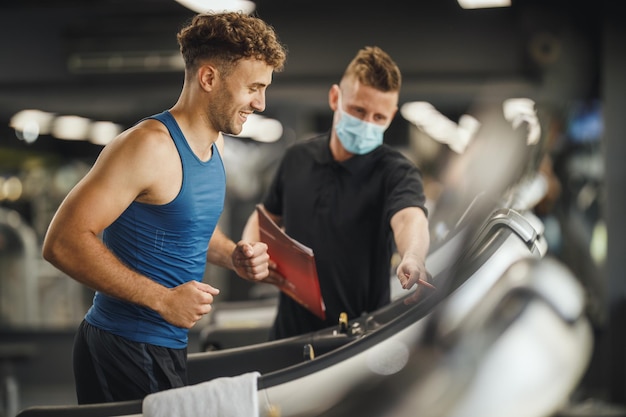 Shot of an muscular young man jogging on the treadmill at the gym. He is working out with personal trainer.