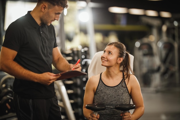 Shot of a muscular young woman in sportswear working out with personal trainer at the gym machine. She is pumping up her muscule with dumbbell.