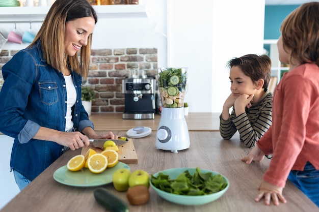 Shot of pretty young woman cutting an apple for preparing detox beverage while her sons look her in the kitchen.