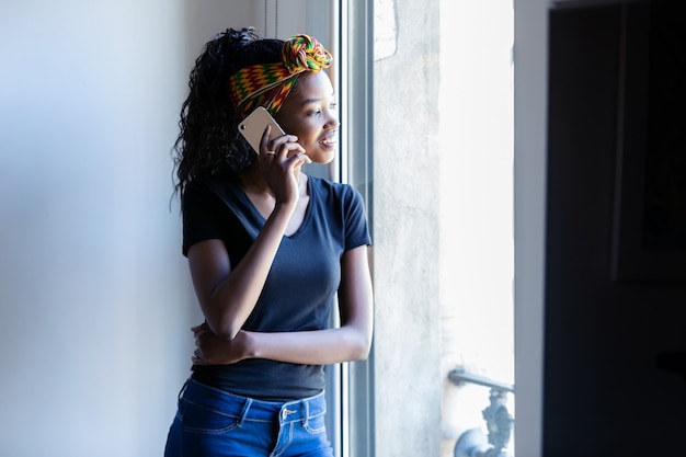 Shot of pretty young woman talking on mobile phone while looking through the window at home.