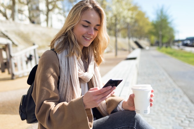 Shot of pretty young woman using her mobile phone while holding cup of coffee in the street.