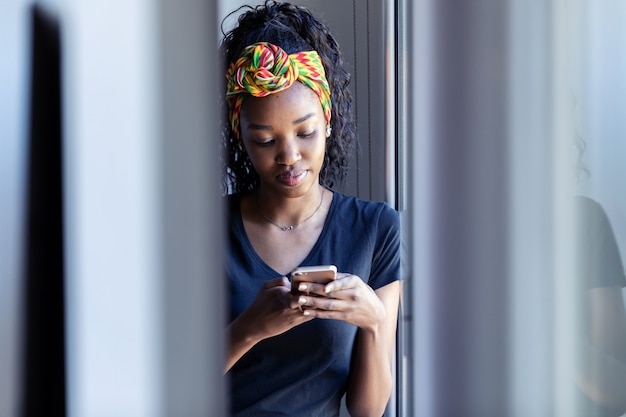 Shot of pretty young woman using her mobile phone while standing next to the window at home.