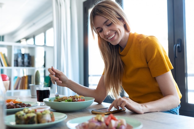 Shot of smiling woman eating healthy food while using her mobile phone at home.