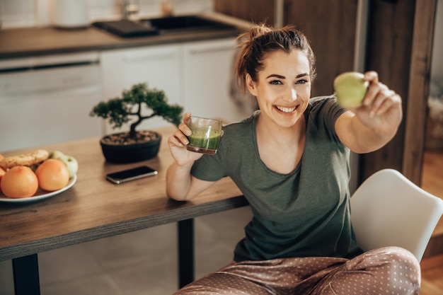 Shot of a smiling woman holding a glass of smoothie and green apple at home in the morning. Looking at camera.