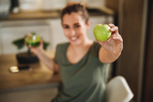 Shot of a smiling woman holding a green apple and glass of smoothie. Selective focus on an apple.