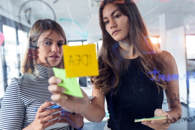 Shot of two business young women working together on wall glass with post it stickers in the modern startup office.