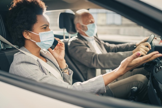 Shot of two successful multi-ethnic business people with protective mask traveling by car during COVID-19 pandemic.