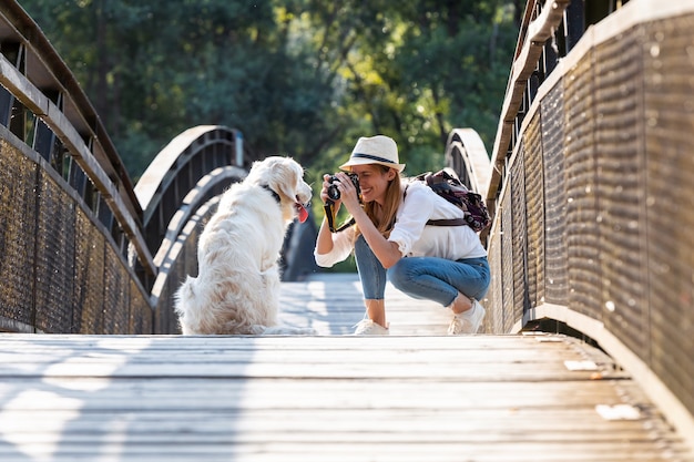 Shot of young amateur photograph woman taking a photo of her dog sitting on a bridge in the park.