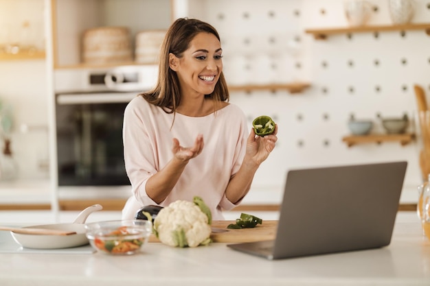 Shot of a young smiling woman preparing a healthy meal and using laptop to make a video blog at home.