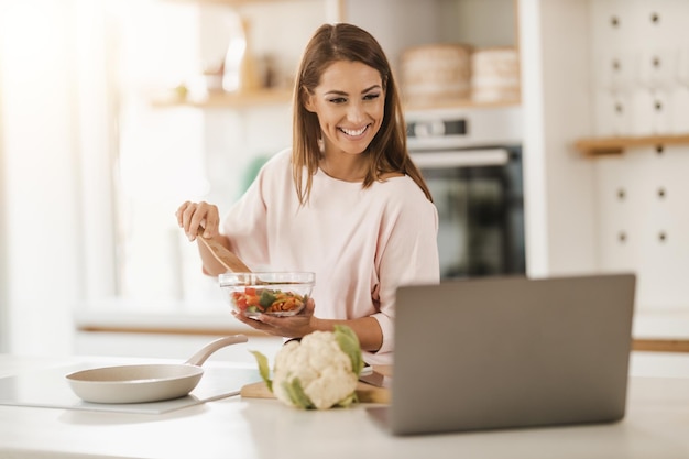 Shot of a young smiling woman using laptop to make a video call with someone while preparing a healthy meal at home.