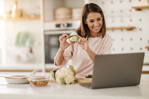 Shot of a young smiling woman using laptop to make a video chat with someone while preparing a healthy meal at home.