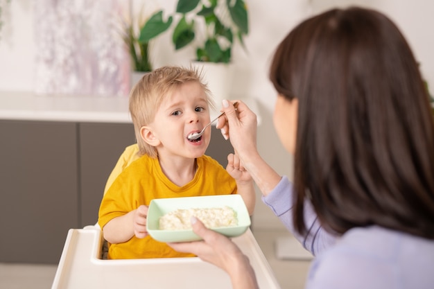 Over shoulder view of mother feeding son with porridge using spoon in kitchen