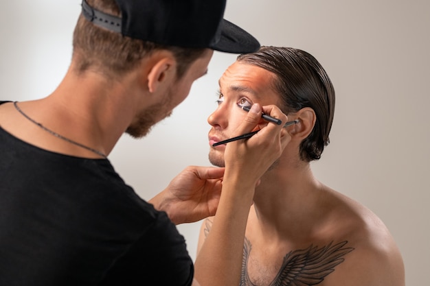 Over shoulder view of MUA artist in cap applying eyeliner to young bearded actor with tattoo on chest in studio