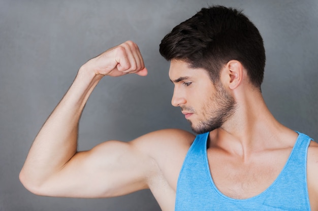 Showing his perfect bicep. Confident young muscular man posing while standing against grey background