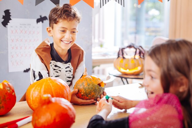 Showing pumpkin. Happy beaming boy wearing skeleton costume showing little pumpkin his beautiful dark-haired friend