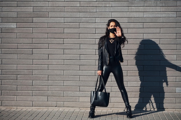 Showing stop gesture by hand In protective mask Beautiful brunette with curly hair and in black clothes outdoors near wall