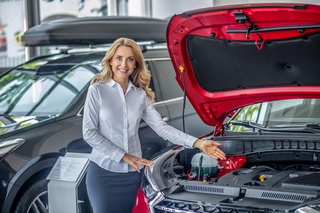 Showroom. Pretty blonde woman standing nea the red car and smiling