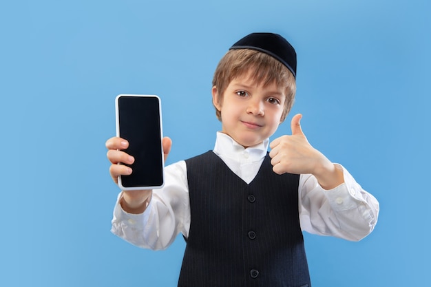 Shows blank phone screen. Portrait of orthodox jewish boy isolated on blue studio wall.