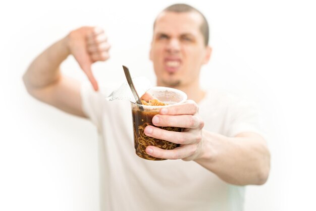 shows thumb down sign and holds a fast food noodles transparent plastic cup on a white background