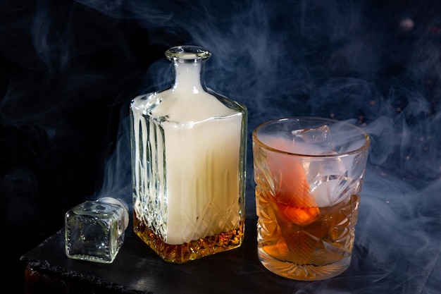 Shrouded in smoke whisky glass decanter with smoke and glass of whiskey with an ice and orange peel on slate plate