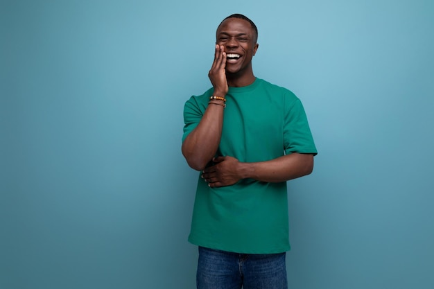 Shy young american man in a tshirt holds his hand to his face on a blue background with copy space