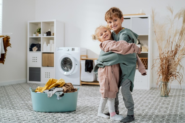 Siblings are standing in middle of the laundry room bathroom next to a bowl of clothes