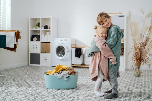 Siblings are standing in middle of the laundry room bathroom next to a bowl of clothes