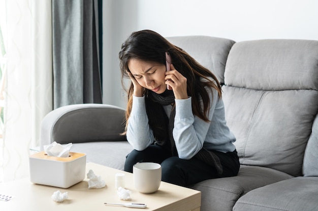 Sick Asian woman sitting on sofa in living room at home and talking with doctor or her family on mobile phone for counseling Cold flu home isolation during coronavirus pandemic concept Closeup