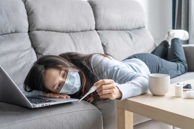 Sick Asian woman wearing medical mask working on laptop from home office. Sad girl with cold lying on sofa while looks at the thermometer. Work from home, health problems and home isolation concept