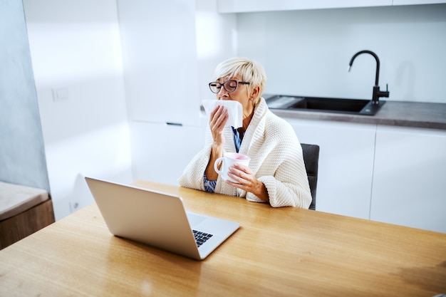 Sick blond senior woman covered with blanket sitting at dining table and sneezing