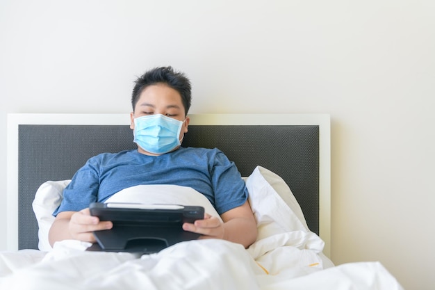 Sick boy wearing mask and studying online with tablet pn bed new normal and education