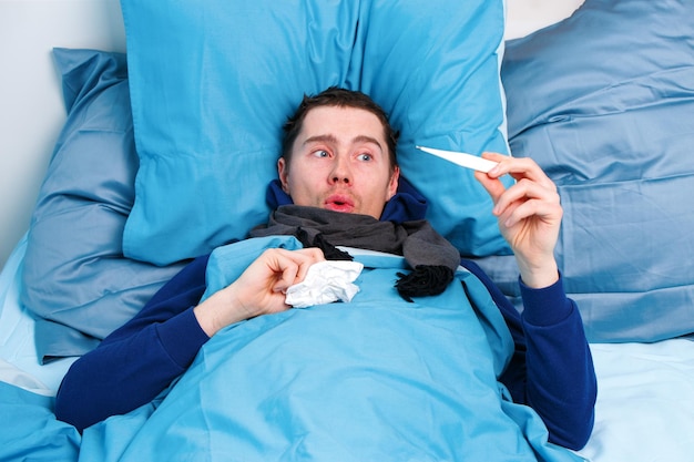 Sick brunet man in scarf with thermometer in hand lying in bed